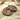 Two people holding wine glasses sit beside a round gift box filled with assorted chocolates and candies, tied with an orange ribbon, on a cream-colored fabric.