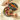 A round gift box with a floral design and orange ribbon, filled with assorted colorful chocolates, sits open on a silky cream-colored fabric background.
